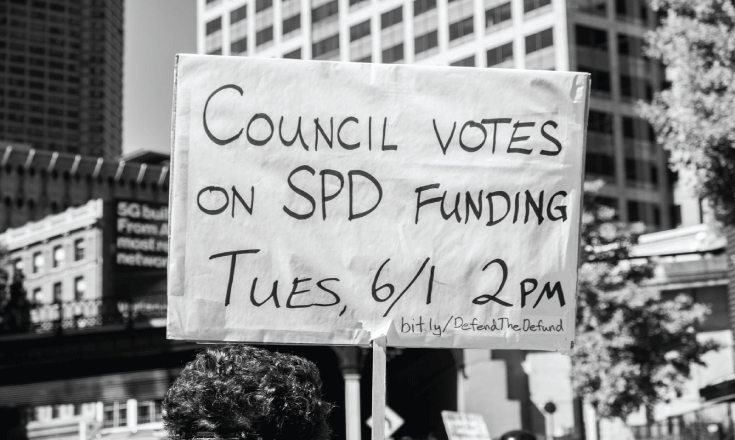 A protestor carries a sign noting a significant Seattle city council vote that would provide an additional $10.9 million in SPD funding